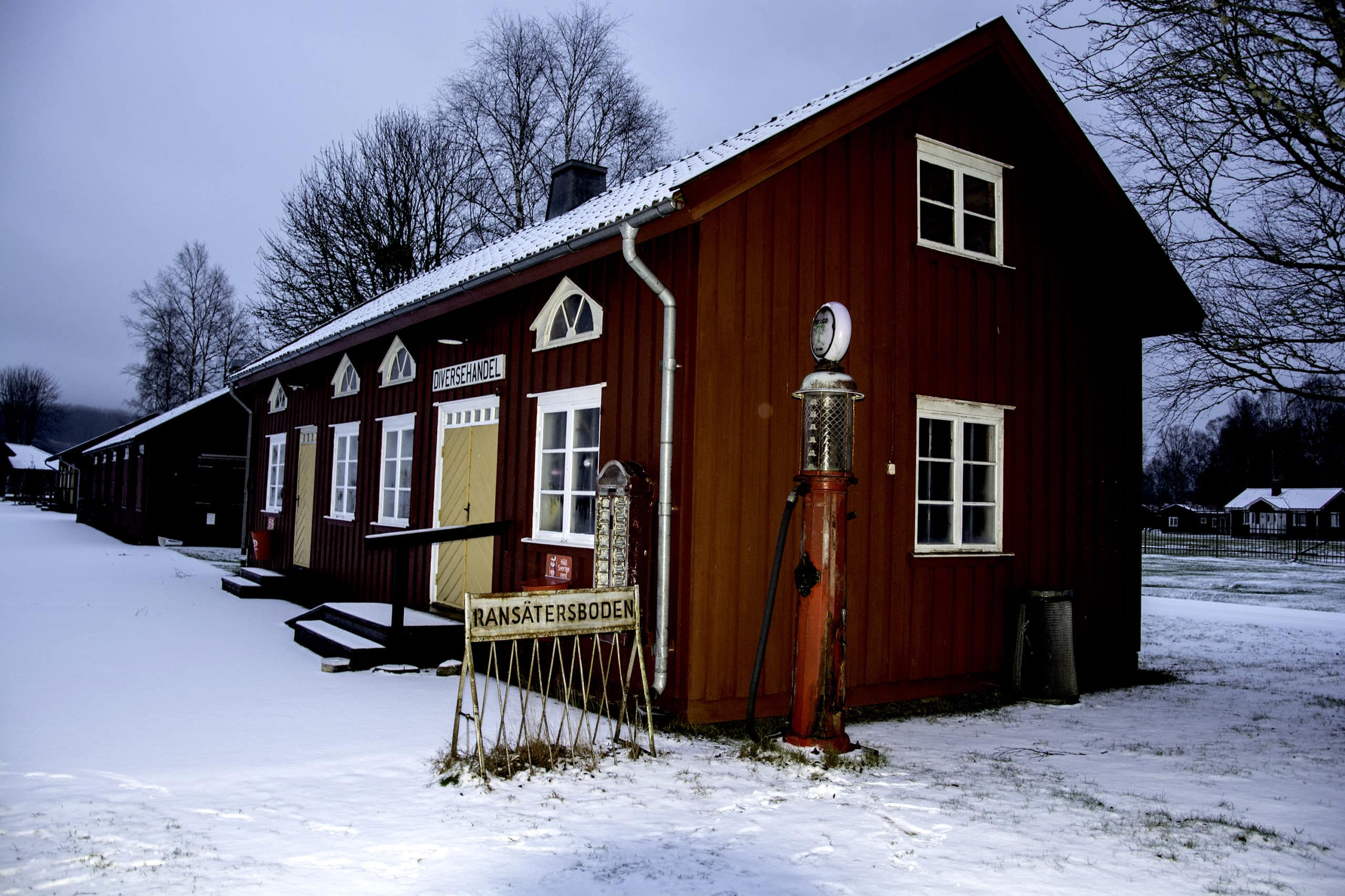 Den röda handelsboden med gul dörr och snö på marken på Ransäters Hembygdsgård.