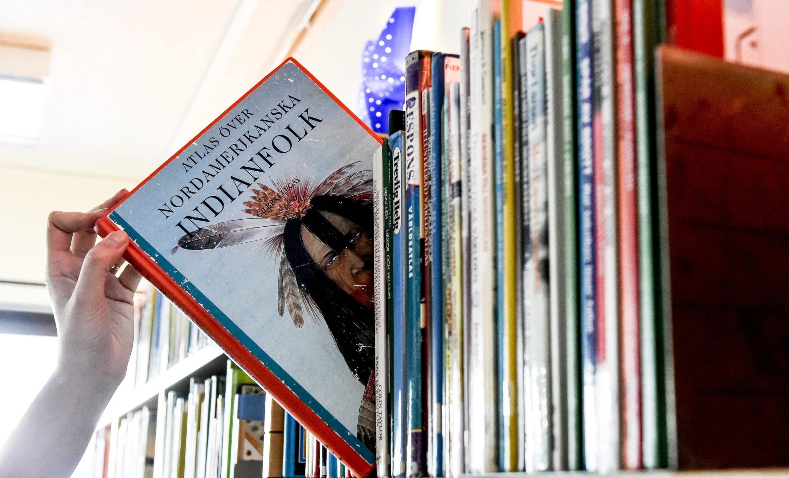 En hand plockar ut en bok "Atlas över nordamerikanska Indianfolk" ur en bokhylla full med böcker på Munkfors bibliotek.