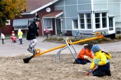 Tre barn i färgglada kläder leker tillsammans i en sandlåda. Två av barnen leker i sanden med varsin plastspade. Den tredje balanserar på en gul gungbräda.