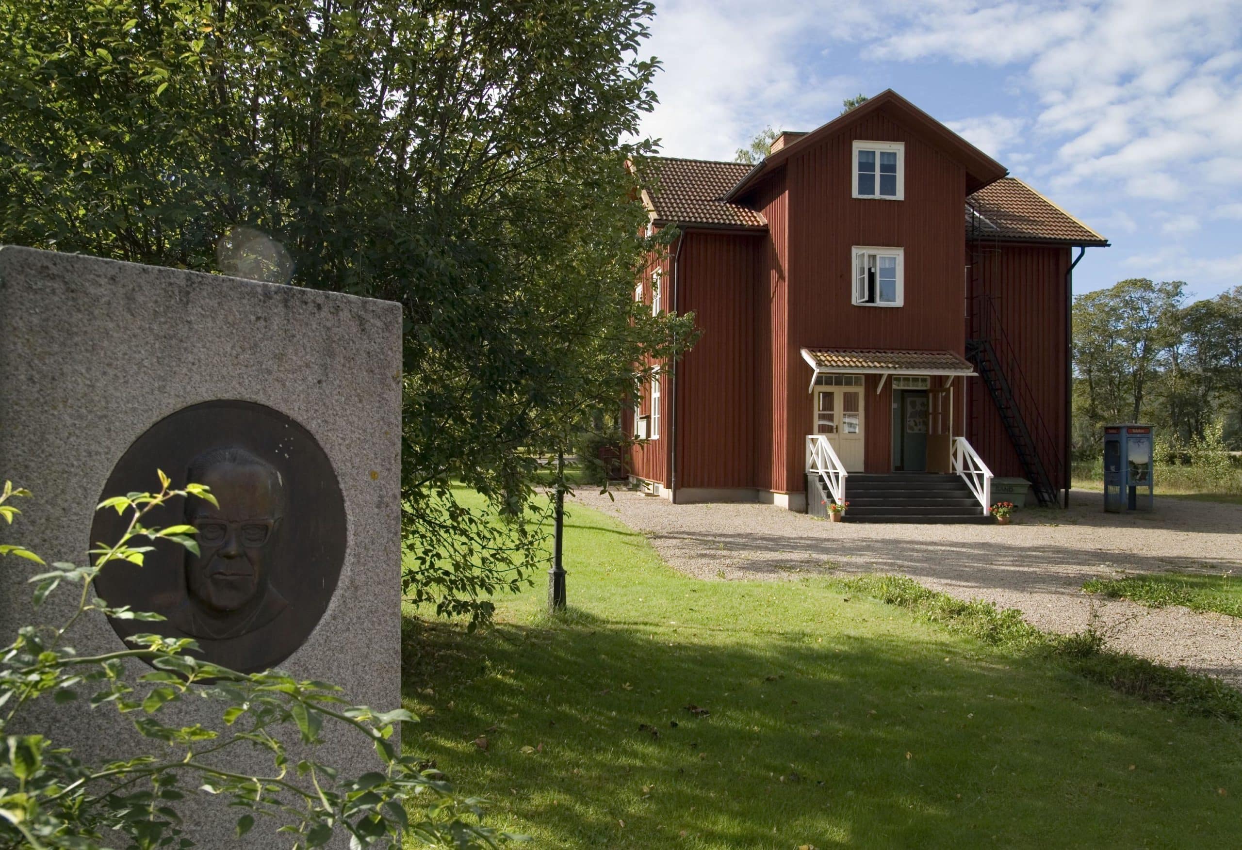 Erlandergården med dess röda fasad och i förgrunden en fyrkantig sten med en bild av Tage Erlander på.