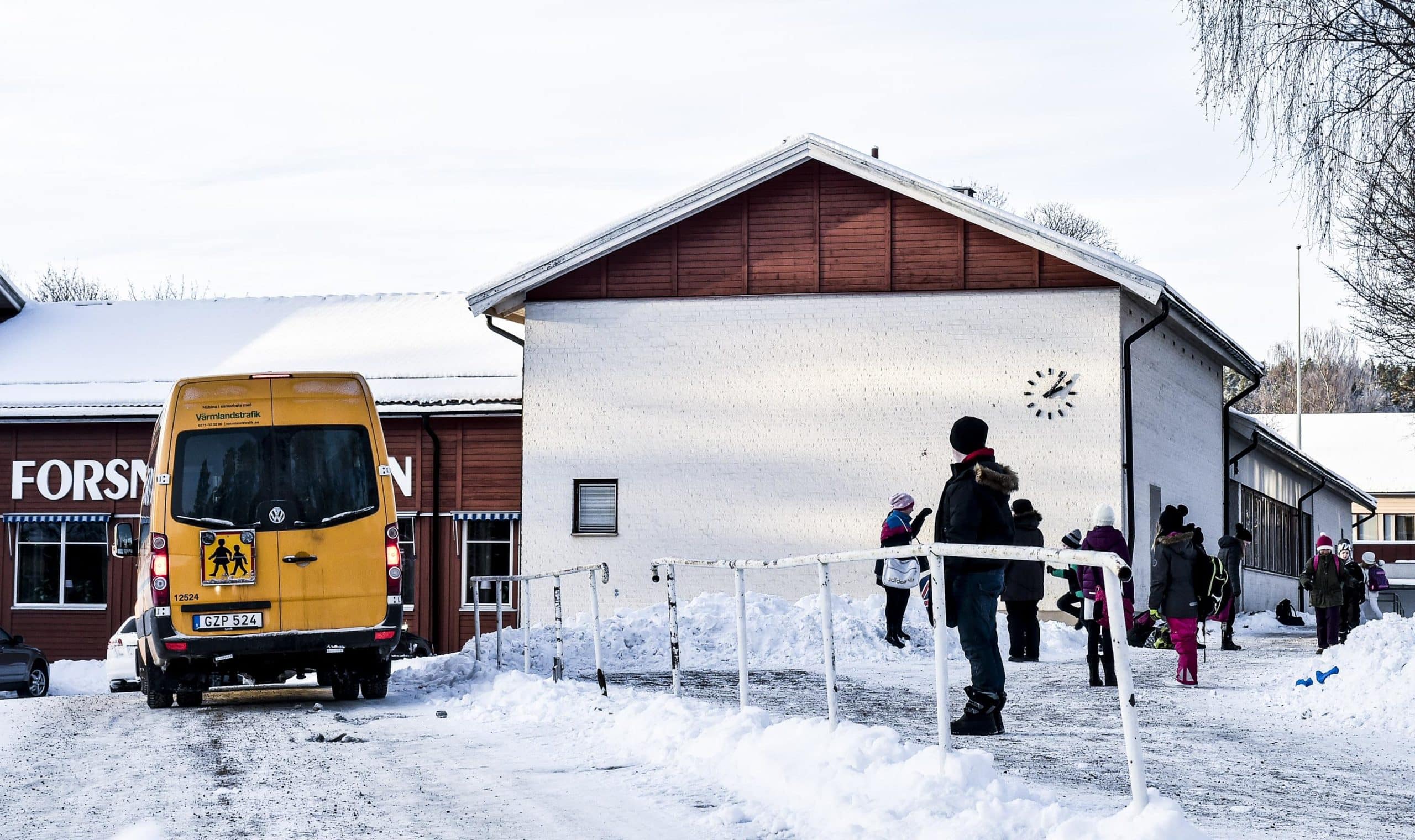 Nio elever i mörka vinterkläder väntar på skolskjuts utanför Forsnässkolan. En liten gul buss passerar framför barnen.