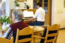 En äldre person i en rödbrun väst sitter vid ett runt bord. Framför den personen sitter en yngre person i en vit skjorta vid ett piano. Båda personerna sitter med ryggen mot kameran.