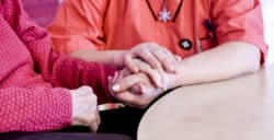 En medarbetare och vårdtagare sitter tillsammans vid ett bord och håller varandras händer.