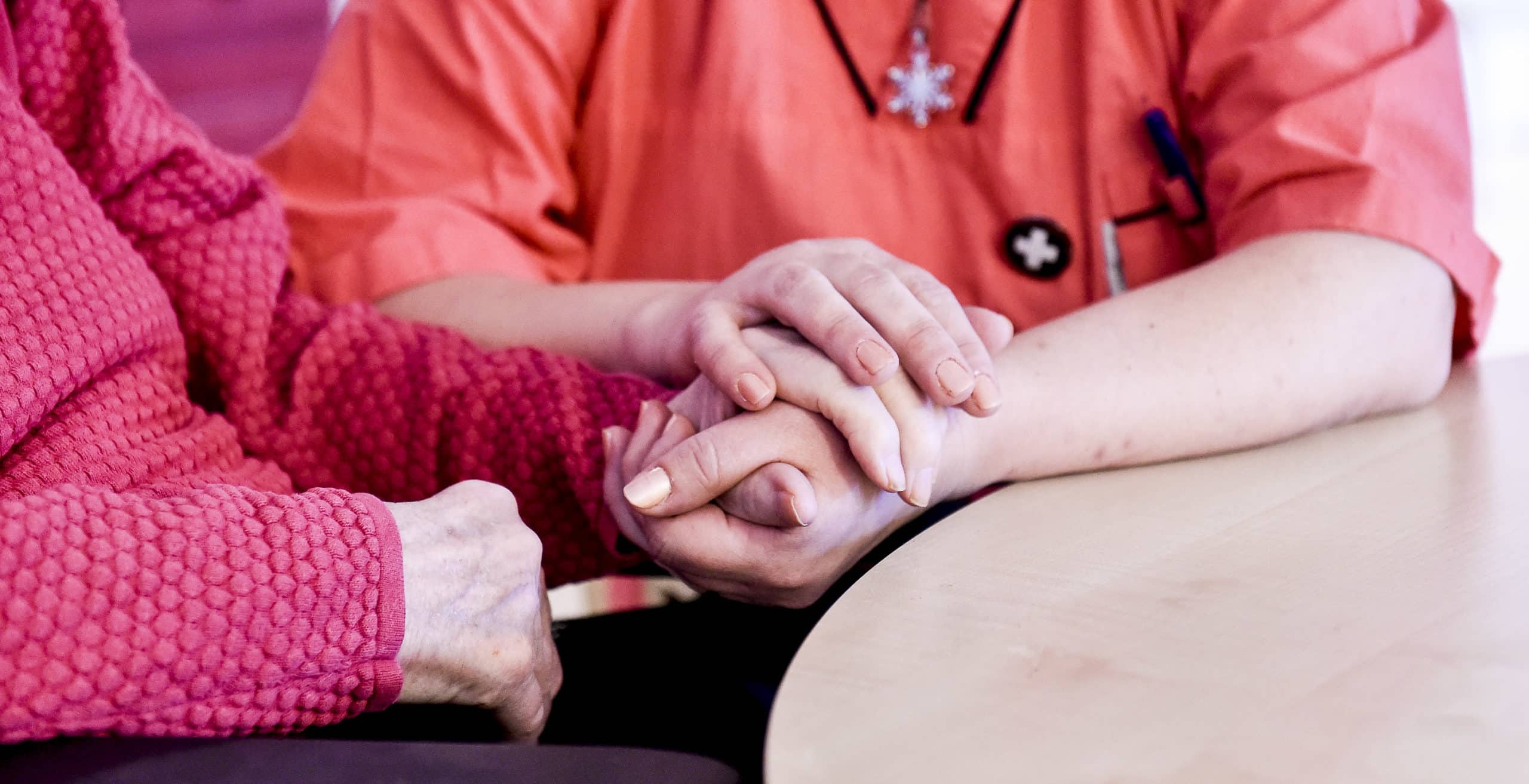 En medarbetare och vårdtagare sitter tillsammans vid ett bord och håller varandras händer.