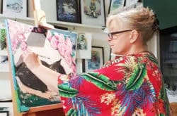 Mia Älegård målar en tavla i sin ateljé. Hon bär en röd kimono med mycket färgglada mönster.
