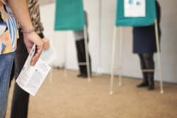 I förgrunden ses en person som står med ett röstkort i handen. I bakgrunden syns två personer som röstar bakom skärmar.