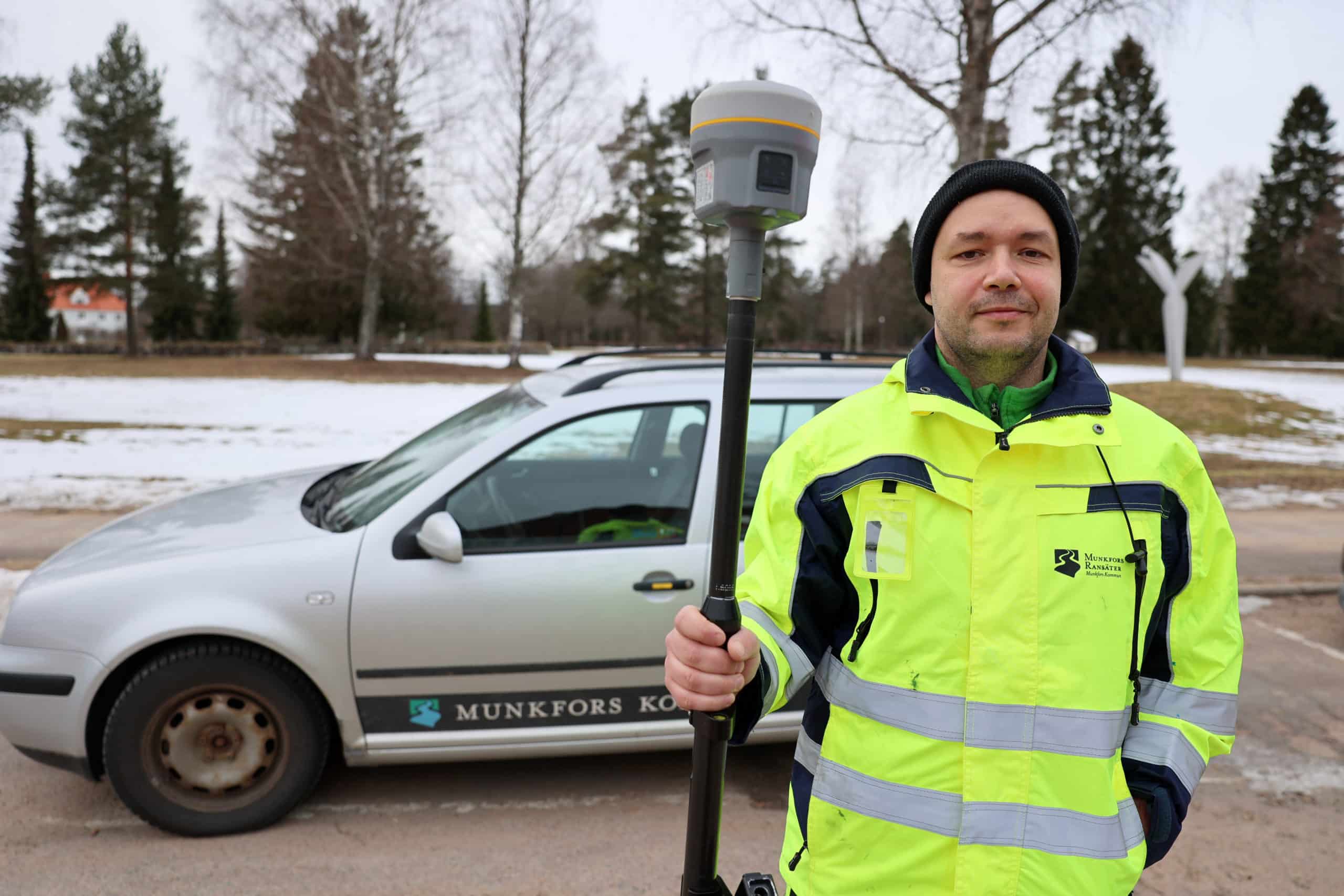 Person håller i ett mätinstrument. Bil med kommunlogotyp är i bakgrunden.