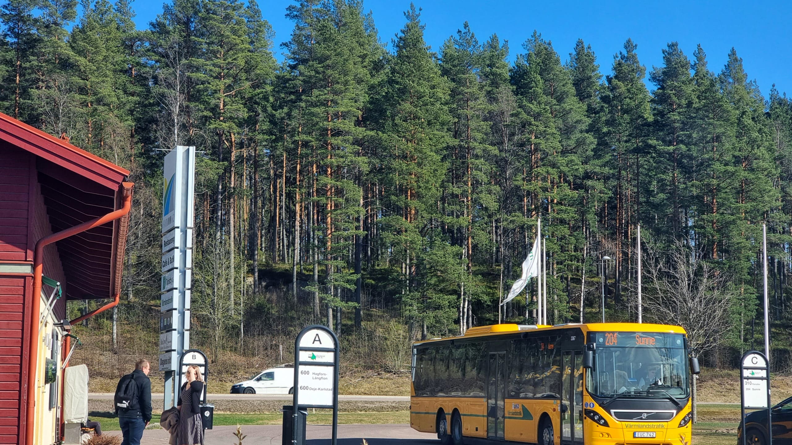 Två personer står vid en busshållsplats. En buss står parkerad med texten Sunne.